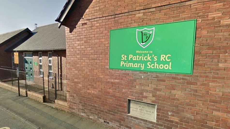 An external picture of St Patrick's Primary School. A large green sign displaying the school's emblem and name is hung on the red brick building, which also contains an inset stone with writing carved into it. The entrance is to the right behind a small metal fence.