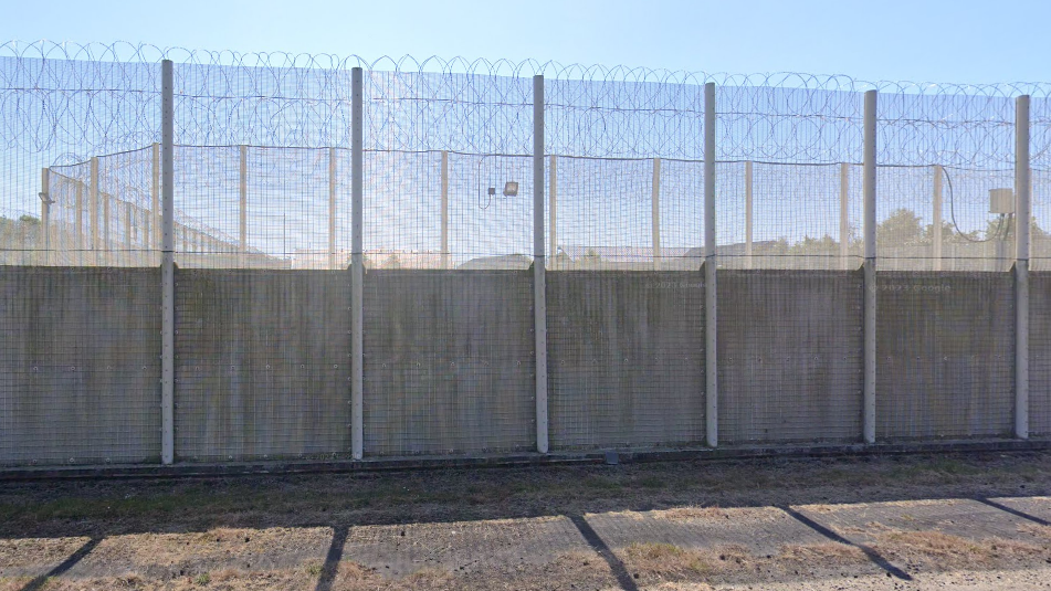 HMP Northumberland exterior. Grey gates around the prison complex. The bootom half of the wall is solid concrete, the top half is made of barbed wire.