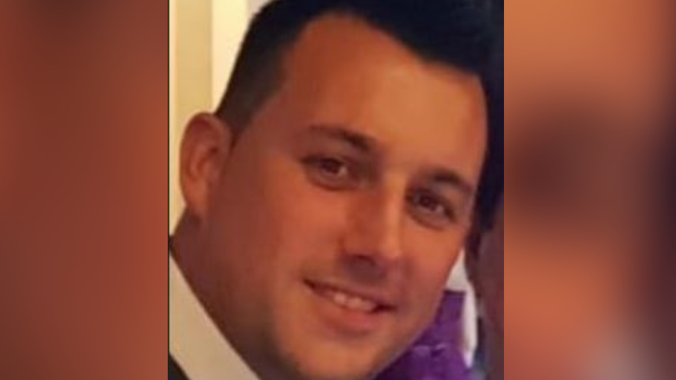 Close up image of Christian Dean smiling. He is wearing a white shirt and has short black hair. The sides of the image are blurred.
