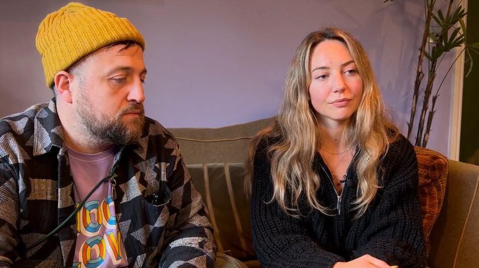 Saf Suleyman, a man with a beard and wearing a yellow beanie hat, and Rebecca Dimmock, a woman with long blonde hair and wearing a black jumper, sat on a sofa.