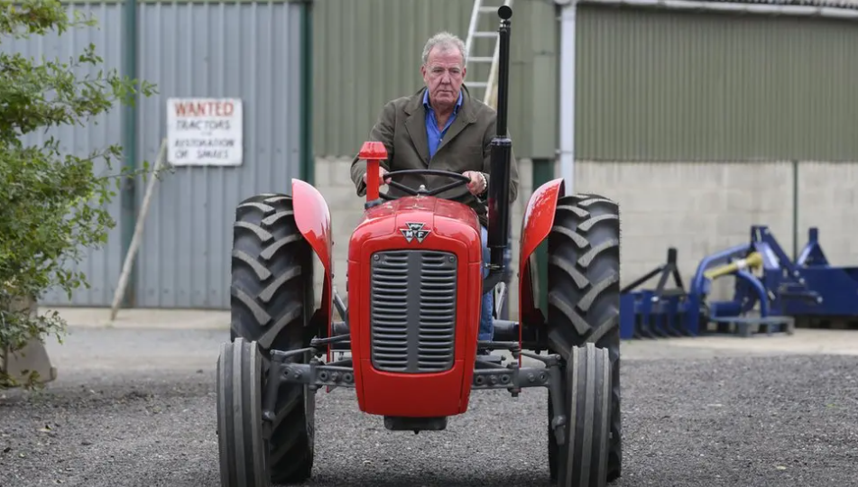 Jeremy Clarkson is sitting driving a red tractor in a green coat and jeans.