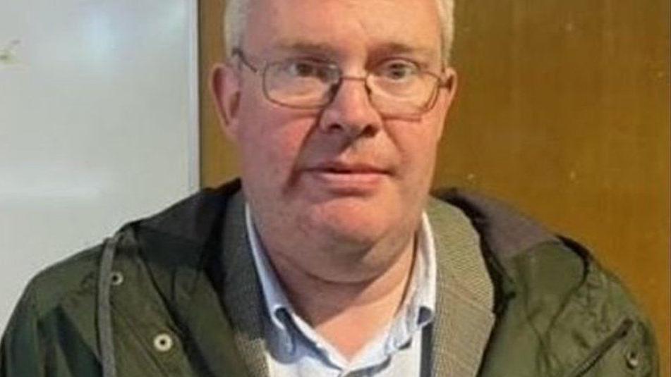 James Raeburn looking into the camera while wearing glasses and a green jacket and blue shirt 