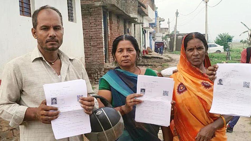 A man in a white shirt, and two women wearing blue-green and orange sarees hold up their enumeration forms during the special roll revision exercise in Bihar, India.