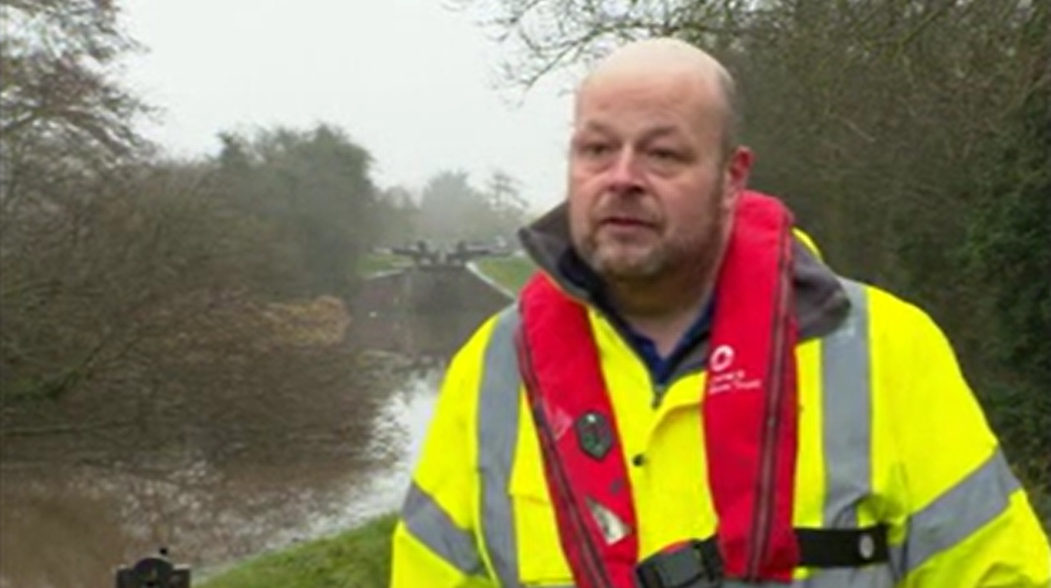 A man with very little hair and a beard stands by the side of a canal. He is wearing a yellow hi-vis jacket and a red flotation device.