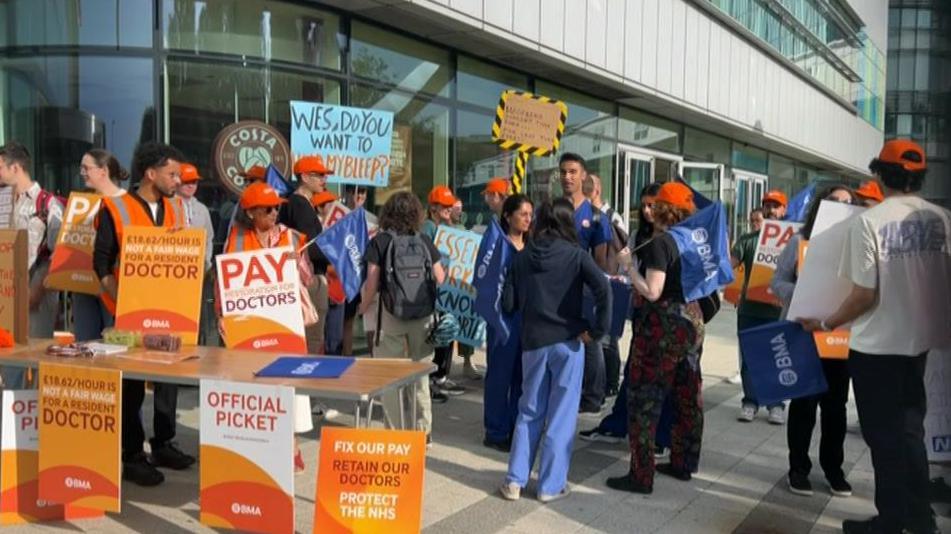 Dozens of people stand outside a hospital building holding placards and signs that say 'official picket' and wearing orange hats.