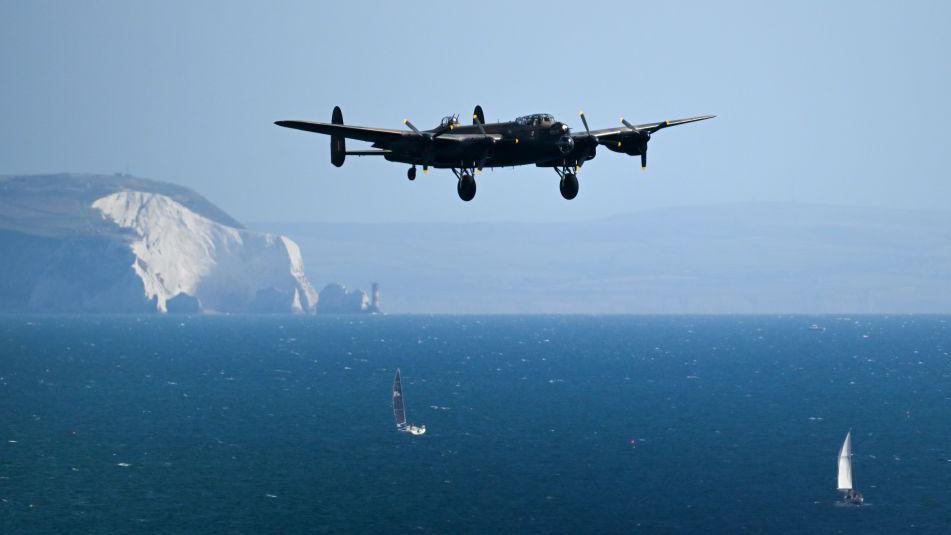 A World War Two RAF Lancaster Bomber in flight over the sea, with the Isle of Wight in the background. Two sailing boats can be seen on the sea.