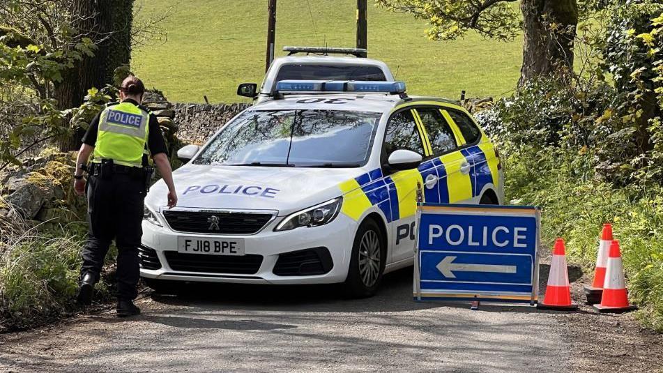 A police officer at road closure in Whaley Bridge in Derbyshire. There are three traffic cones, a police sign, a stationary police car and an officer on a narrow country lane.  