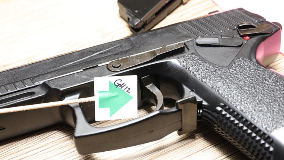 A close up of a black gun on a wooden background. There is a square of paper with a big green arrow symbol attached to a stick, seen by the trigger.