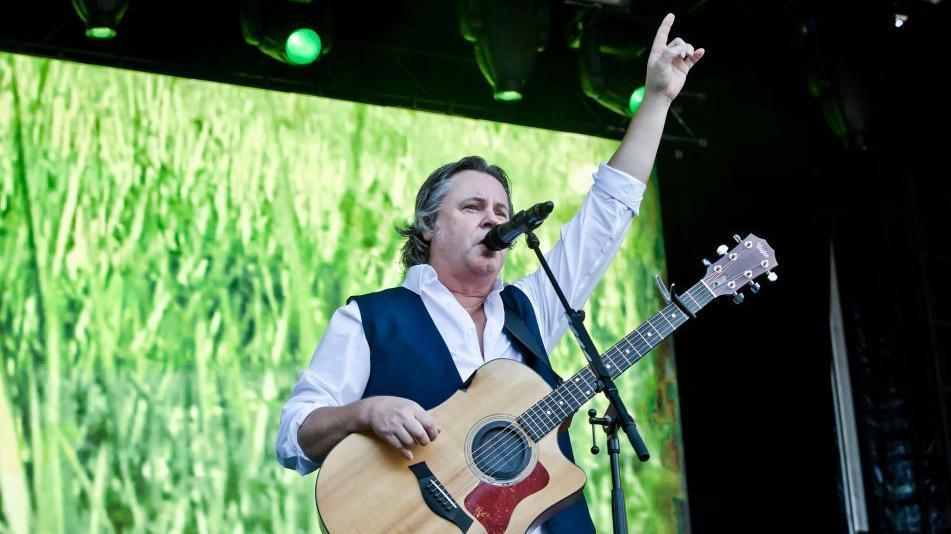 Runrig performing on stage. The singer is wearing a white shirt and a dark waistcoat. He has his arm raised and is singing into a mic. He has a guitar over his shoulder