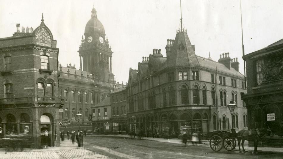 A black and white photograph of Leeds around 1900, with people walking on the streets and a horse and cart in the foreground.