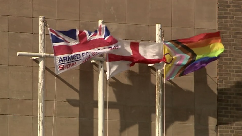 Three flags on white poles outside a building. One is a British Armed Forces Union Jack flag, one is the St George's flag and one is the Progress Pride Flag.