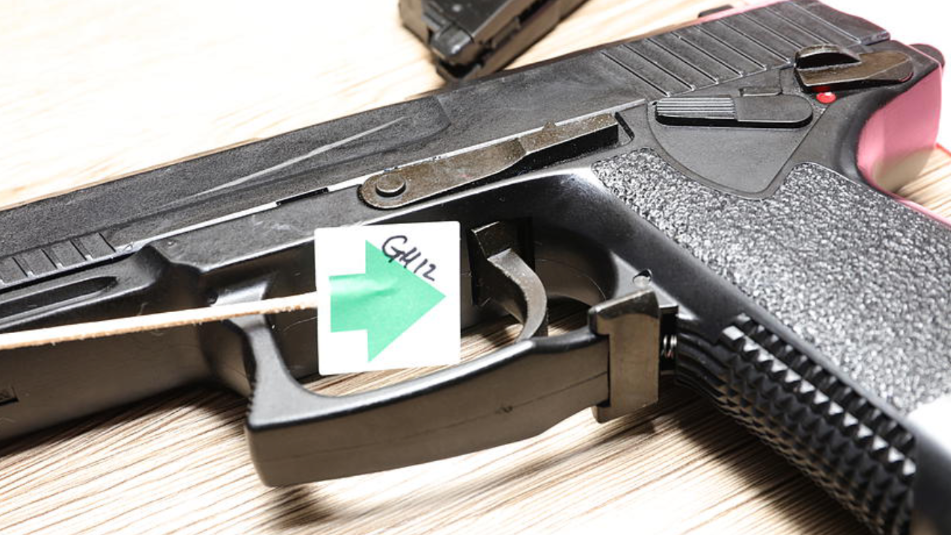 A close up of the one of the hand guns. It has a label with a code and a green arrow pointing at the trigger. The gun in on its side on top of a light wood grain table top.