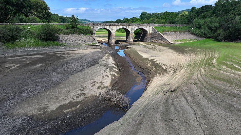 aerial photograph of a water reservoir with a very small amount of water running through a riverbed and a stone bridge in the background - the area looks dry