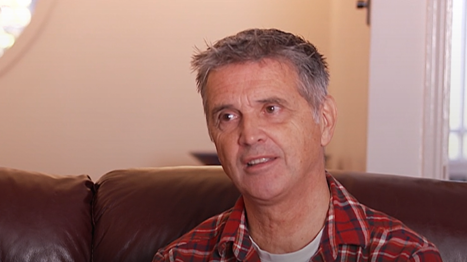 A man with short grey hair wearing a red plaid shirt. He is sitting in a brown leather chair looking to the left of the camera