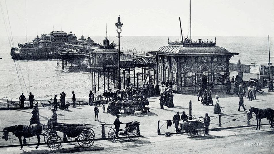 A black and white image of the West Pier. There are horses and carriages in front of the pier, on the road, and groups of people walking. 