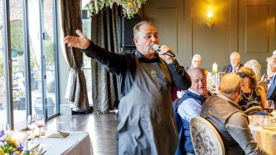 A waiter in a black shirt and dark brown apron holds his hand out while singing into a microphone at a wedding. Guests can be seen sat on tables watching him sing.