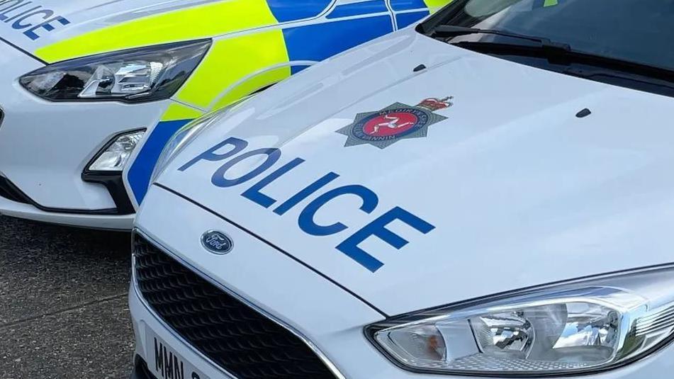 The bonnets of two police cars. The cars are white with blue writing spelling police alongside the Isle of Man Constabulary crest, which is blue and red and features the three legs of Mann. The side panels of one car, which are blue and bright yellow, can be seen.