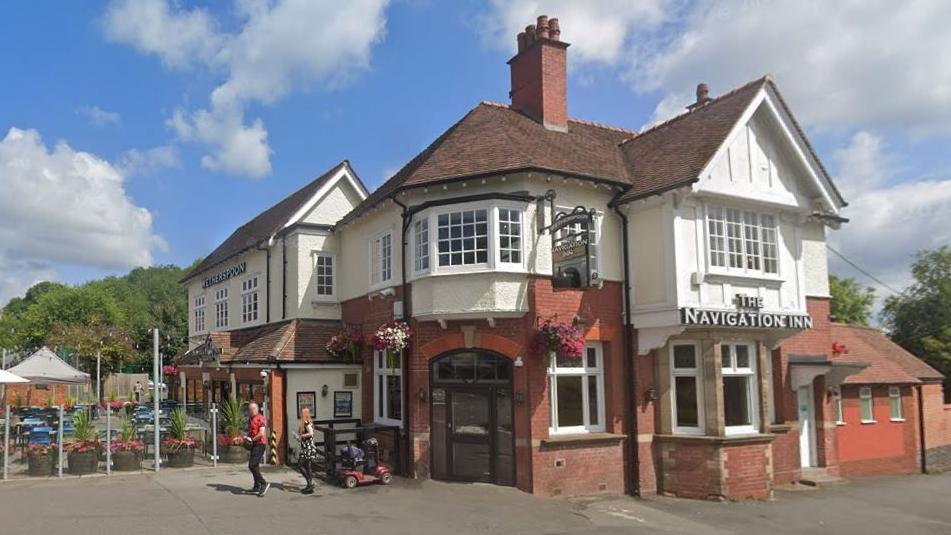 A large redbrick pub building on a sunny day. The building has two storeys and an outdoor beer garden area. A sign on the front of the building says "The Navigation Inn".