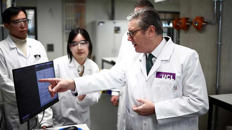 Keir Starmer, in a white lab coat, points at a computer screen, watched by a young man and women in lab coats and safety goggles. He is on a visit to the Manufacturing Futures Lab at University College London