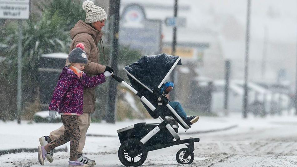 A woman pushes a buggy. A young girl, who is wearing a bright purple coat and grey hat, walks alongside her. They are crossing an icy road as snow continues to fall.