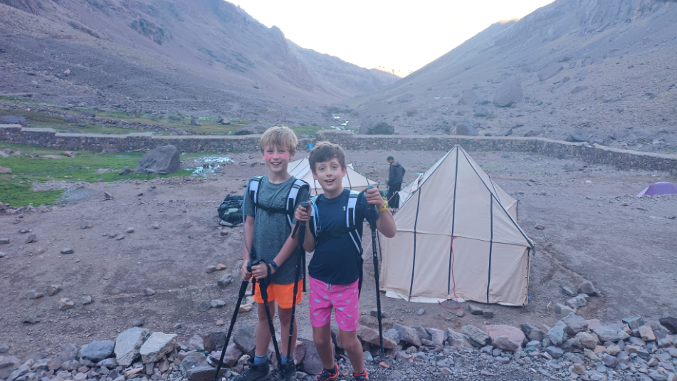 A bleak mountain landscape with Freddie and Max holding walking poles and wearing rucksacks, with two makeshift tents in the background