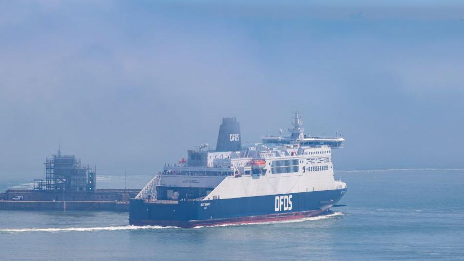 A DFDS ferry on the channel. It is blue and white with the name of the company on the side in large white letters. It is passing a structure in the sea which has a concrete jetty and scaffolding on it.