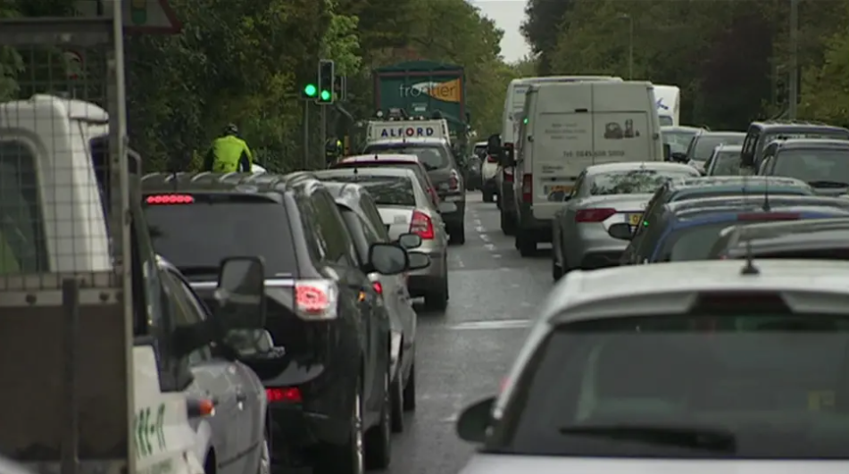 Three lanes of back-to-back traffic heading away from the camera, with green trees wither side of the road.