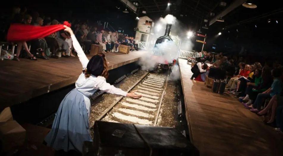 A picture of a steam train on a railway line in a specially adapted theatre with a woman stood in front of it waving a red cloth and audience members sat either side.