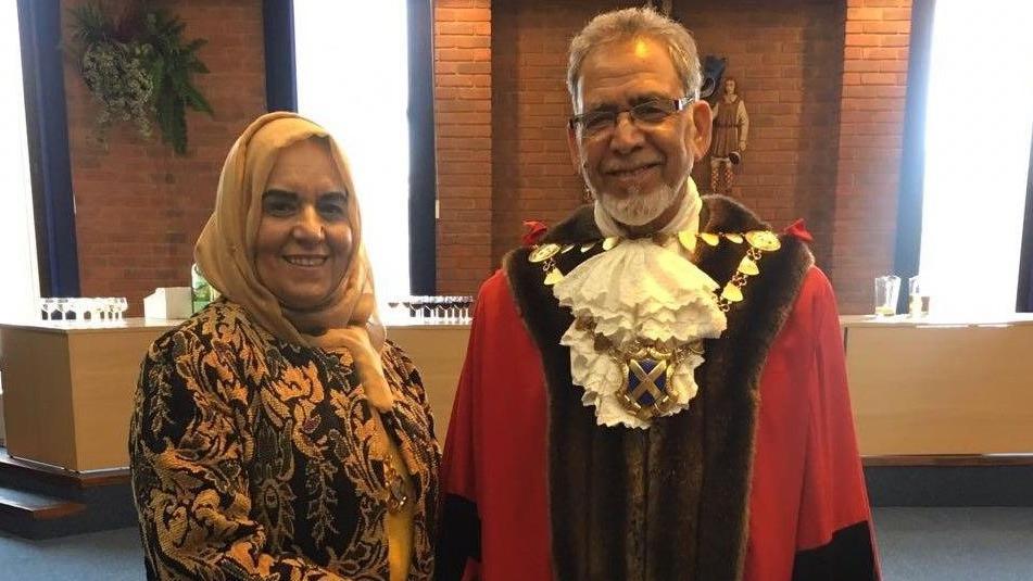 Farhat (left) and Iqbal Zia in a room, both looking at the camera and smiling. Farhat is wearing a cream head scarf and brown and cream patterned jacket. Iqbal is wearing full mayoral robes, with a mayoral chain on, white ruffled shirt, red coat, with brown fur trim. He has glasses on and short grey hair. 