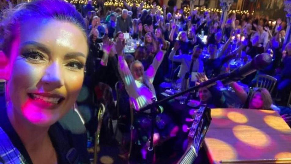 A woman, Kerry, takes a selfie on the stage. There is a crowd of people in the background with their arms raised in the air in joy.