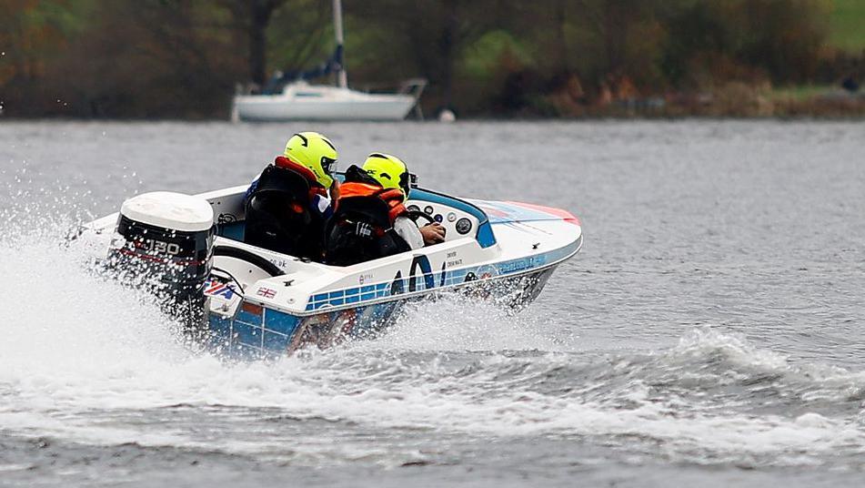 A blue and white power boat on the water moving at speed. There are two people wearing fluorescent helmets in the boat. There is a white boat in the background.