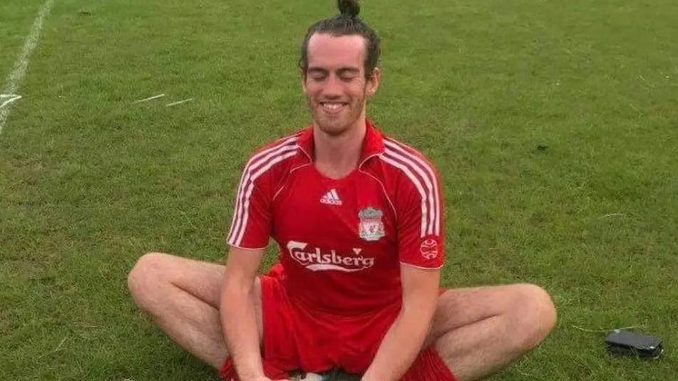 Liam McGenity sat crossed legged on a football pitch wearing a Liverpool football club kit