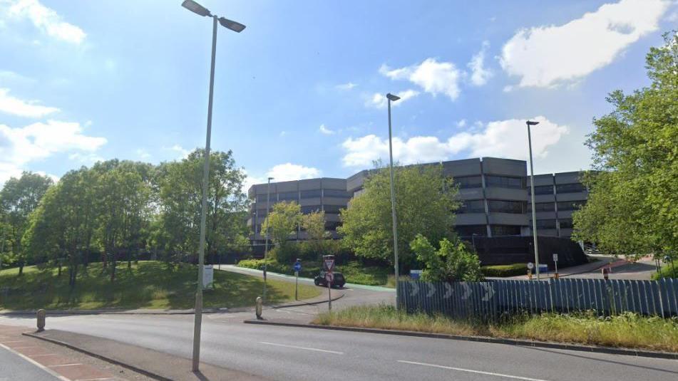 A large grey building, with three four-storey towers visible rises behind large green trees and a grassy area to the left. A two-lane main road runs from left to right in front of the site