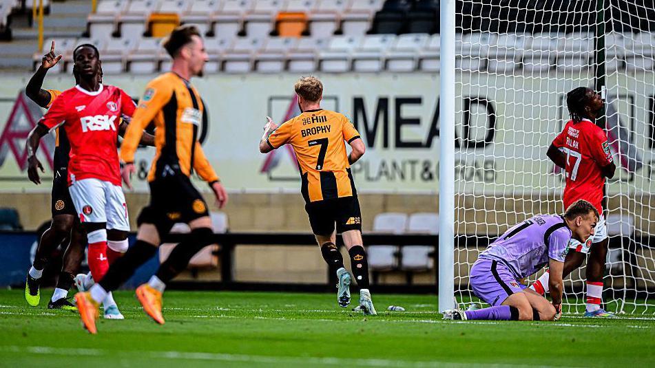 James Brophy celebrates after scoring for Cambridge United, running away from goal pointing towards teammate Elias Kachunga, who set up the goal, as Charlton Athletic players look dejected