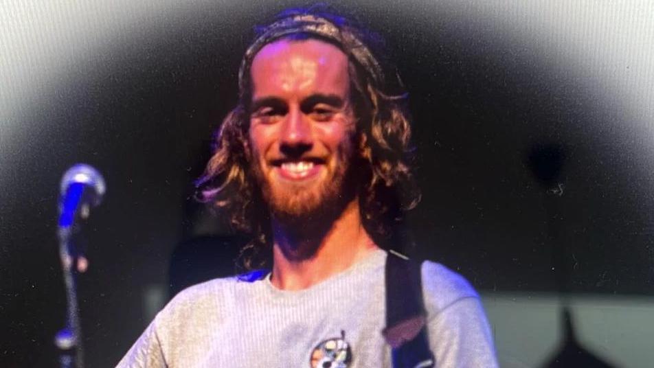 Liam McGenity smiling on stage stood behind a microphone wearing a grey jumper with a guitar strap over his shoulder.