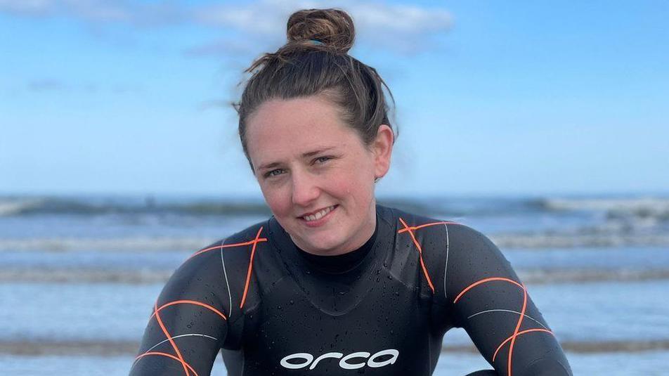 A picture showing Jasmine Harrison who is smiling and wearing a wetsuit. In the background is the sea. 