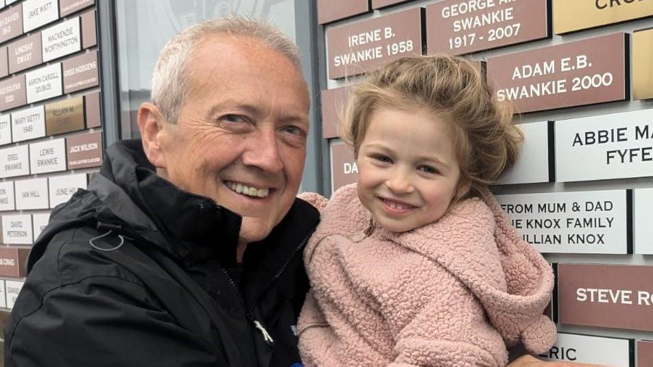 Stan wears a black raincoat, and holds Maggie who is wearing a light pink fluffy zip-up hoodie. They are both smiling at the camera in front of a wall covered in brown, white and gold brick-like plaques with people's names on them