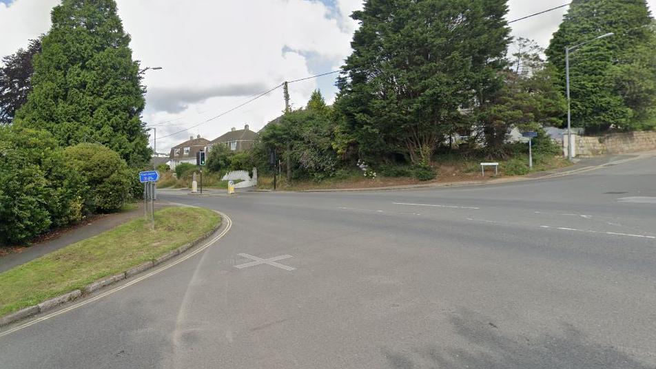 A crossroads junction in a residential area. There is a street name sign at one corner and there is a green verge and tress obscuring houses.