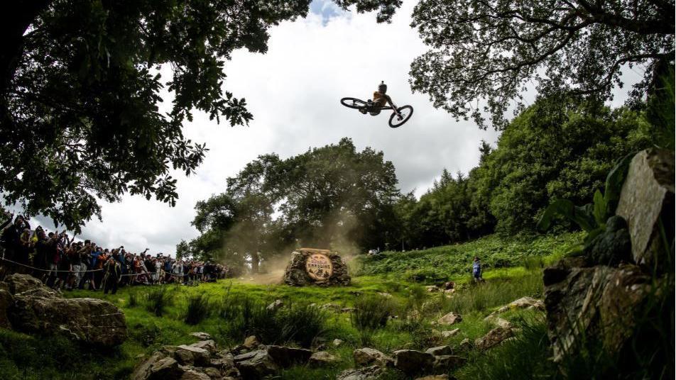 A mountain-biker is in mid-air between trees as people watch on