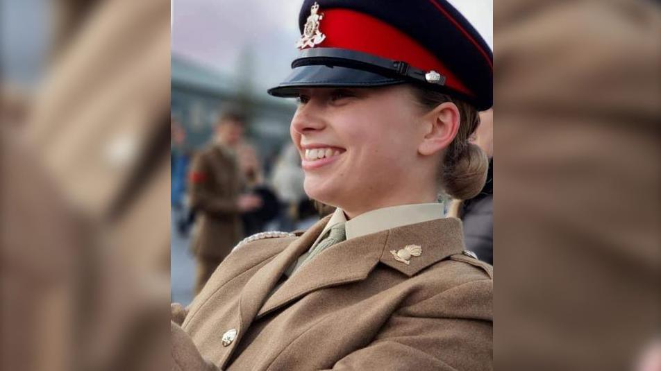 Jaysley Beck wearing her brown formal military uniform and a black and red cap. She has her light brown hair tied back into a low bun and is looking off to the left of the camera and smiling.