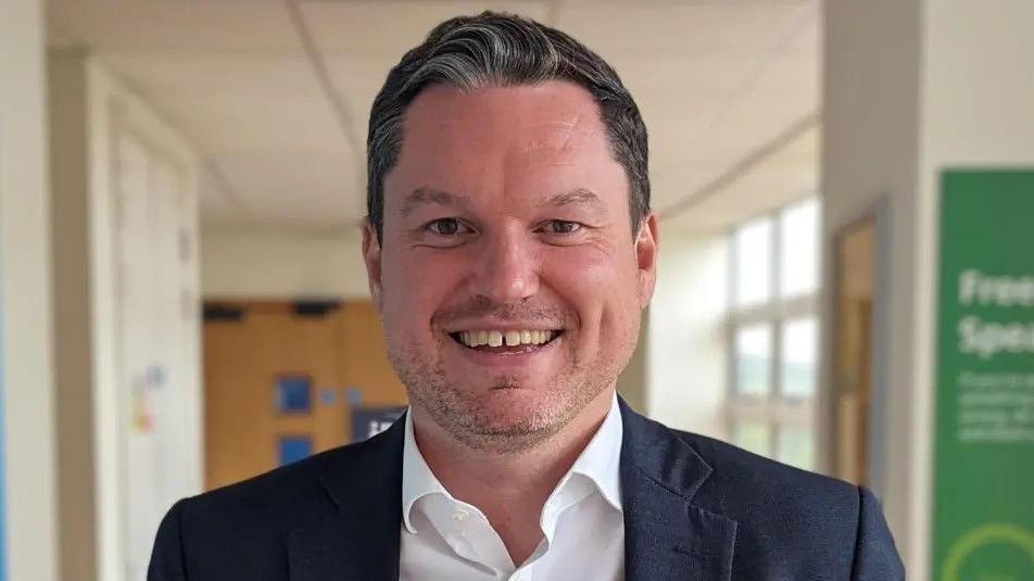 A picture of Kevin McNamara in a corridor, he is wearing a navy blazer and a white shirt, and smiling widely.
