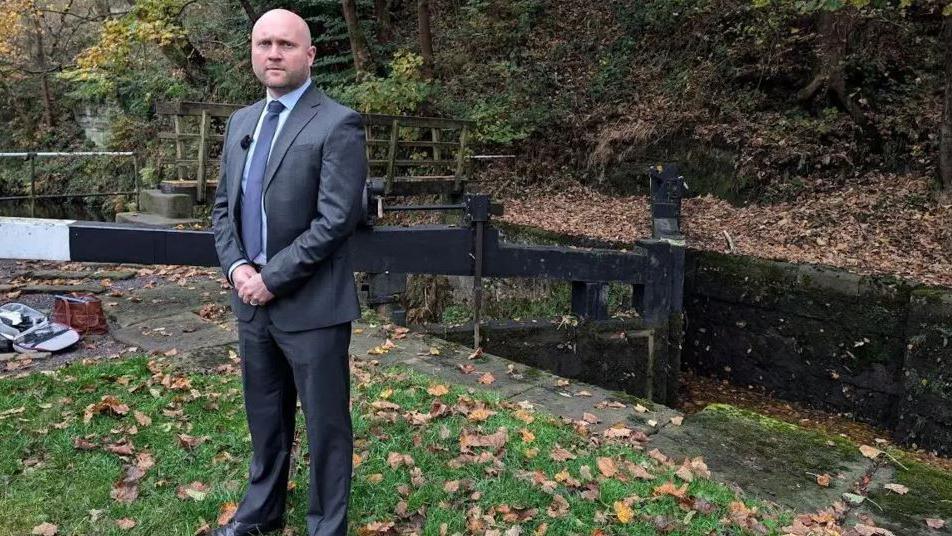 Det Ch Insp James Entwistle, a man in a grey suit and tie, stands next to a canal and there are autumn leaves around him. 