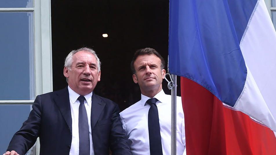 François Bayrou, wearing a suit and tie, stands next to France's president Emmanuel Macron, alongside a French flag