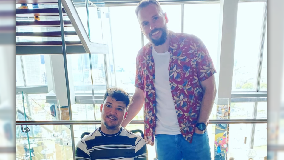 Two men smile at the camera, one is in a wheelchair