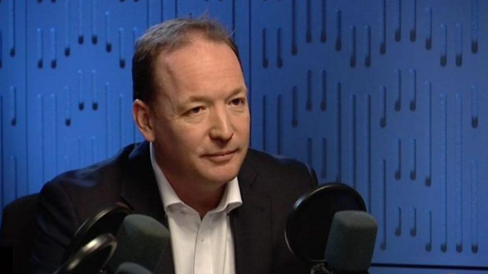 Pete Redfern with short dark hair wearing a black jacket and white shirt sitting in front of microphones in a studio with a blue wall.