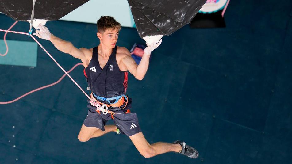 Olympic climber Toby Roberts, wearing his navy Team GB tank top and shorts, hangs from two white holds on a climbing wall, during the Paris 2024 Olympics.