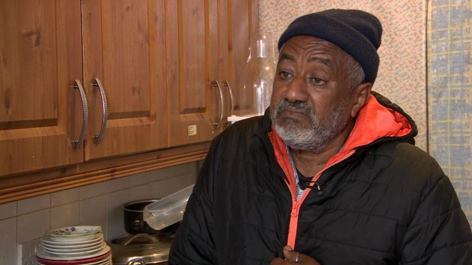 Abdullah Ali wearing a jacket and hat, standing in a kitchen with cabinets behind him