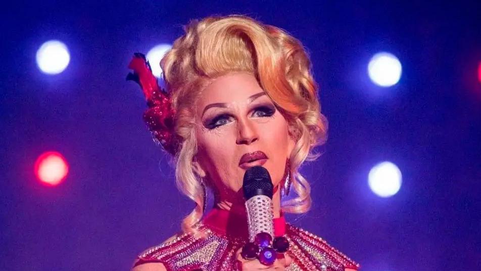 Head and shoulders shot of Zoe in full drag in a blond wig, hair piled high and holding a microphone against a blue background with show spotlights