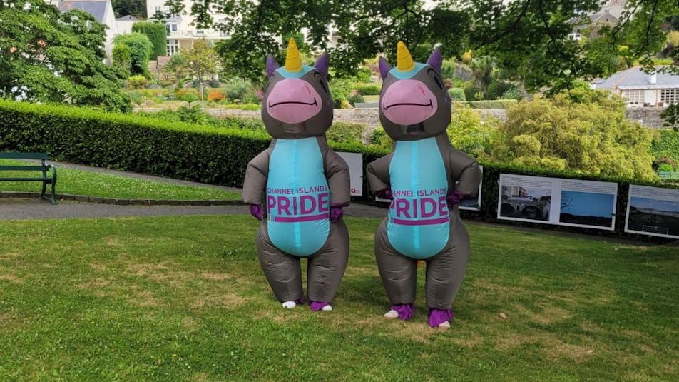 Two inflatable donkey unicorns, with a blue belly, grey skin, pink nose smiling and a yellow unicorn horn, on the stomach saying 'CHANNEL ISLANDS PRIDE' in a garden under a tree in the shade on a sunny day