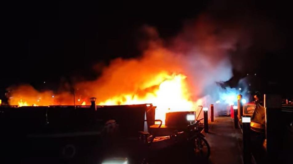 Fire in a marina at night.  Flames are visible surrounded by smoke.  A painted narrowboat name sign is visible to the right. A boat's chimney is visible to the left. 